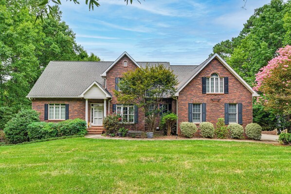 A two-story brick house with a front porch, a well-maintained lawn, and mature trees. - 'sunset Central' - Lake Norman Getaway With Dock (Mooresville)