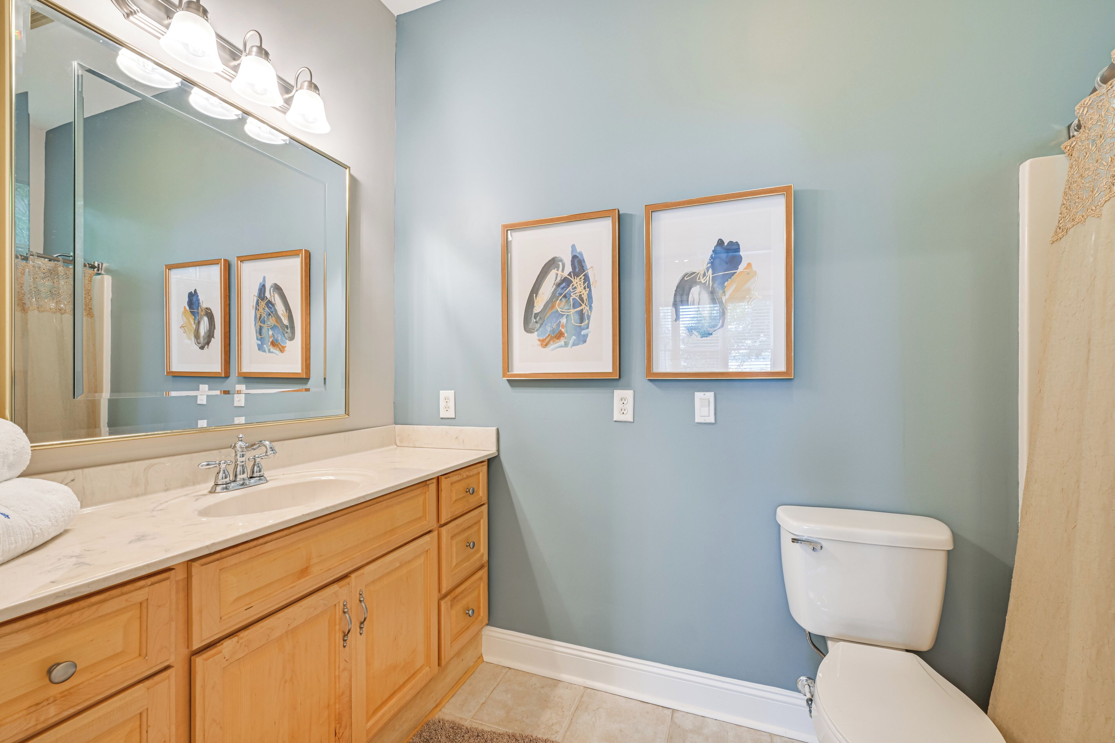 A bathroom with a large mirror, wooden vanity, white sink, framed artwork on the wall, and a toilet.