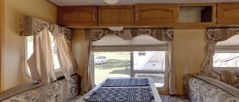 A compact RV interior with a dining table, two cushioned seating areas, and a window providing a view of a residential area.