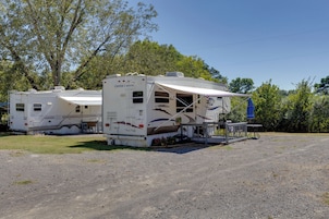 Dos vehículos recreativos estacionados en un camping con suelo de grava, rodeados de árboles.