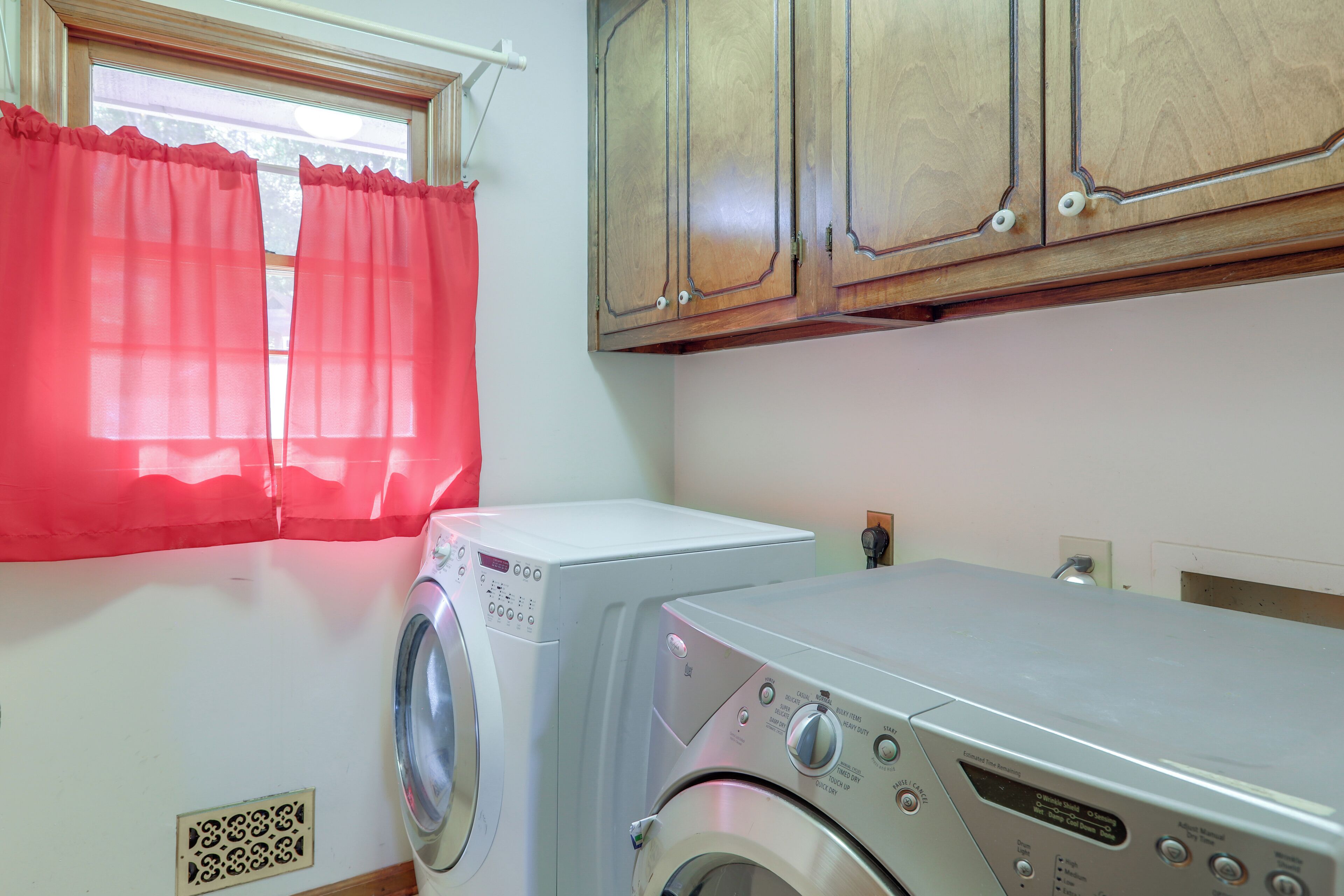 Une buanderie équipée d’un lave-linge et d’un sèche-linge à chargement frontal, de placards en bois au-dessus, et d’une fenêtre avec des rideaux rouges.