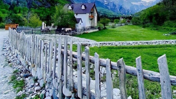 A house with a sloped roof, surrounded by a stone wall and a wooden fence, set against a backdrop of mountains and a clear sky. - Bujtina Tinari (Theth)