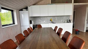 A modern kitchen with white cabinets, a built-in oven, and a dining area with a long wooden table and leather chairs. - 5 Star Holiday Home in Vaeggerlose (Vaeggerlose)