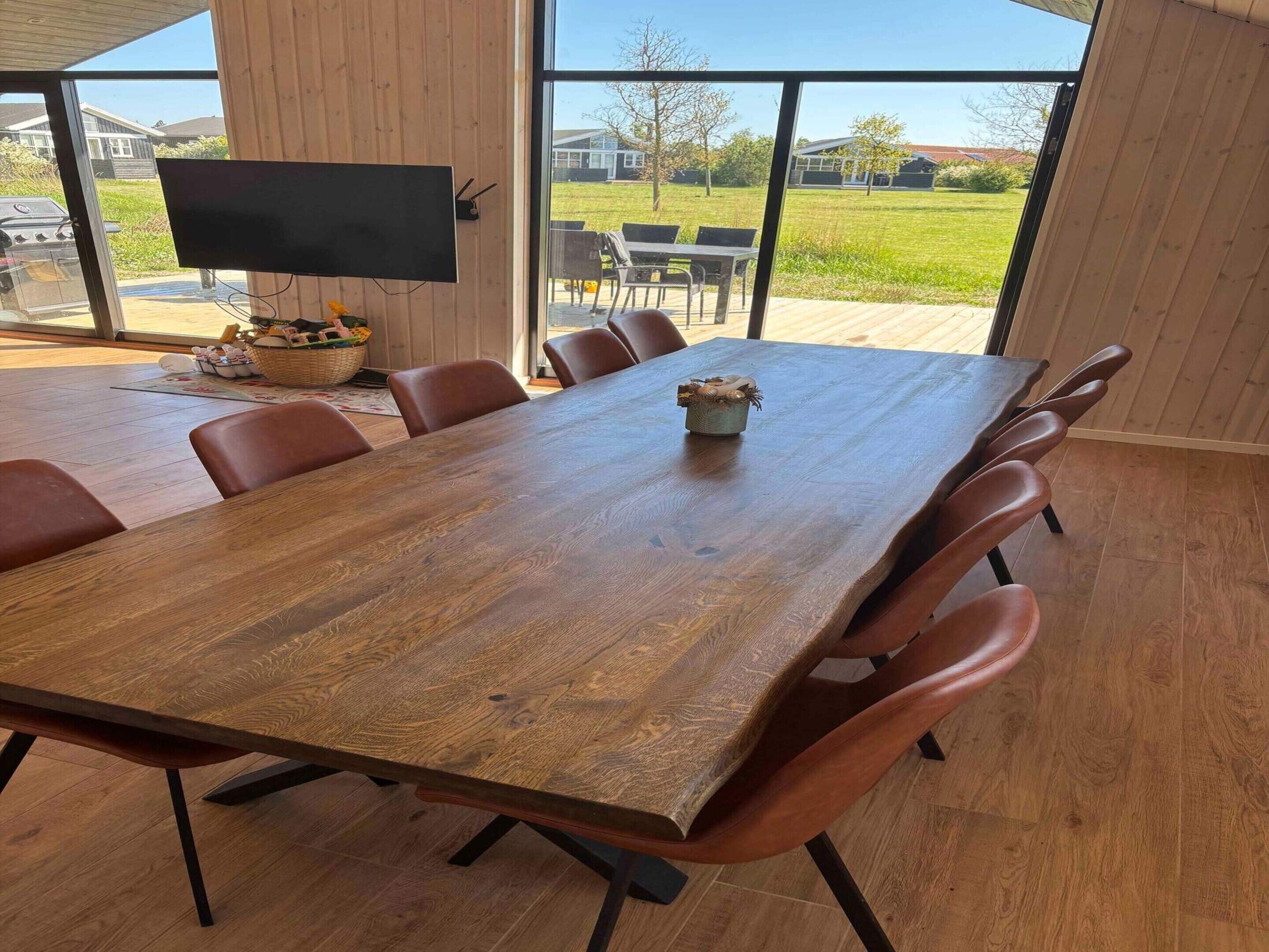 A large wooden dining table with six chairs, a television on the wall, and a view of a grassy area outside.