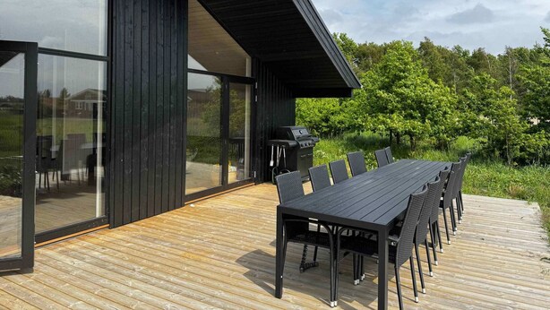 A modern outdoor dining area with a long table and chairs, a grill, and a glass-enclosed space. - 5 Star Holiday Home in Vaeggerlose (Vaeggerlose)