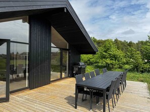 A modern outdoor dining area with a long table and chairs, a grill, and a glass-enclosed space. - 5 Star Holiday Home in Vaeggerlose (Vaeggerlose)