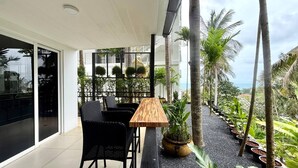 A balcony with a wooden table and black wicker chairs, surrounded by potted plants and palm trees, overlooking a lush landscape. - B11 Sunset Plaza Partial Seaview 1 Bedroom 1 Bathroom With Large Jaguzzi (Karon)