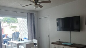 A room with a ceiling fan, a TV mounted on the wall, a dining table with chairs, and a wooden cabinet. - Pelican Perch - Cozy Retreat, steps from the Ocean (Kure Beach)
