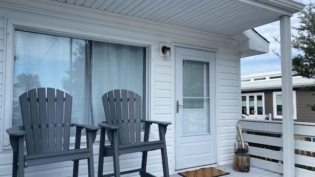 A porch with two Adirondack chairs, a white door, and a small wooden bucket. - Pelican Perch - Cozy Retreat, steps from the Ocean (Kure Beach)