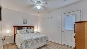 A bedroom with a bed, bedside lamps, a ceiling fan, a door with a window, and a small table with a lamp. - Pelican Perch - Cozy Retreat, steps from the Ocean (Kure Beach)