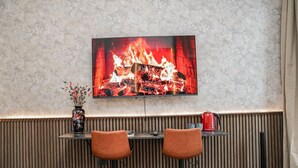 Ein moderner Barbereich mit einem Fernseher, der einen Kamin zeigt, zwei braune Ledersitze, einer Vase mit Blumen und einem roten Wasserkocher auf einem Regal.