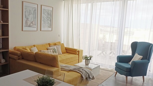 A living room with a yellow sofa, a blue armchair, a coffee table, and a bookshelf. - Near to Victoria. Cozy 2 bedroms penthouse with Citadel view. (Ta' Sannat)