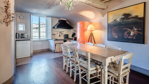 Un coin repas avec une table et des chaises en bois, un lustre, une cuisine équipée d’une cuisinière et de placards, et un grand tableau accroché au mur.