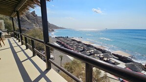 A beach view with a balcony overlooking the ocean, a sandy beach with umbrellas, and a rocky cliff in the background. - Classic Quadruple Room 1 with Balcony and Air Conditioning (Perissa)