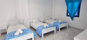 Une chambre avec trois lits simples, chacun recouvert dâune couvre-lit Ă motifs bleus et agrĂ©mentĂ© dâoreillers blancs, et une fenĂȘtre ornĂ©e dâun rideau bleu.
