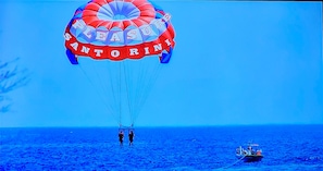 Un parachute ascensionnel sur lequel on peut lire les mots « Pleasure Santorini », deux personnes debout en dessous, et un bateau sur lâeau.