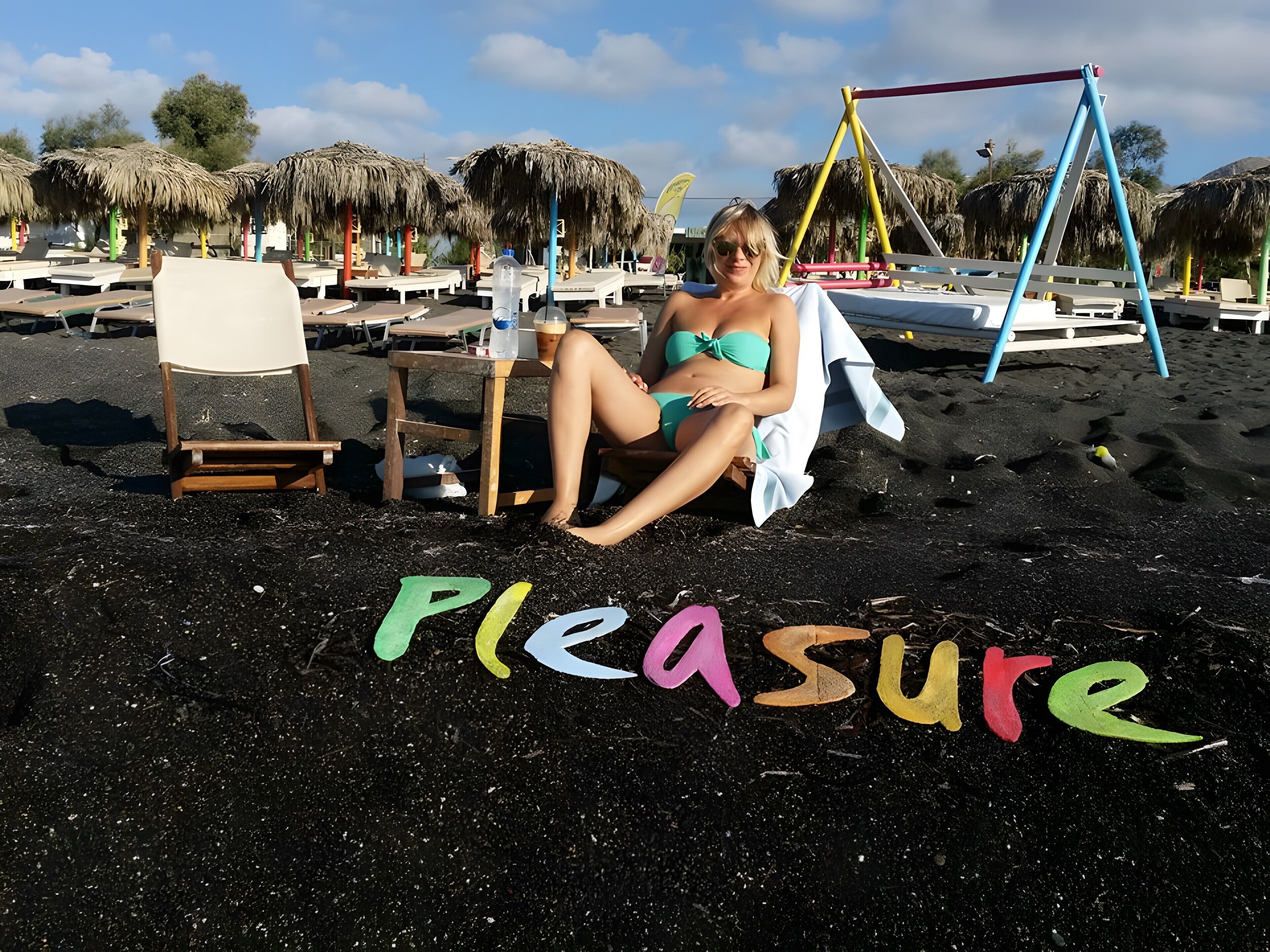 A beach scene with a person sitting on a chair, a swing set, and the word 'pleasure' painted on the sand.