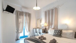A hotel room with a bed, a desk, a chair, a TV, and a lamp. - Double Room with View 8: Sea View, Balcony & Air Conditioning (Perissa)