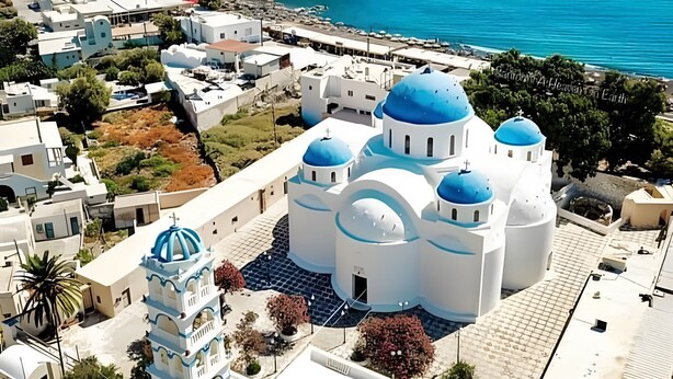 Una cittadina costiera con una chiesa bianca imponente dalle cupole blu, un alto minareto e una scogliera rocciosa sullo sfondo.