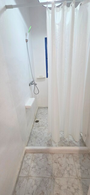A shower area with a white curtain, a handheld showerhead, and a small shelf with a soap dish. - Economy Double Room 3 with Balcony and Air Conditioning (Perissa)