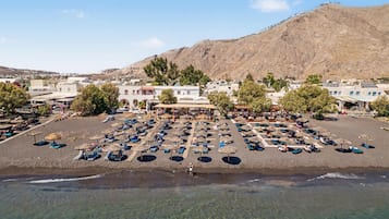 A beachside resort with sun loungers and umbrellas, a central building with a roof, and a mountainous backdrop.
