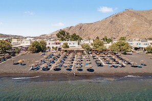 A beachside resort with sun loungers and umbrellas, a central building with a roof, and a mountainous backdrop.