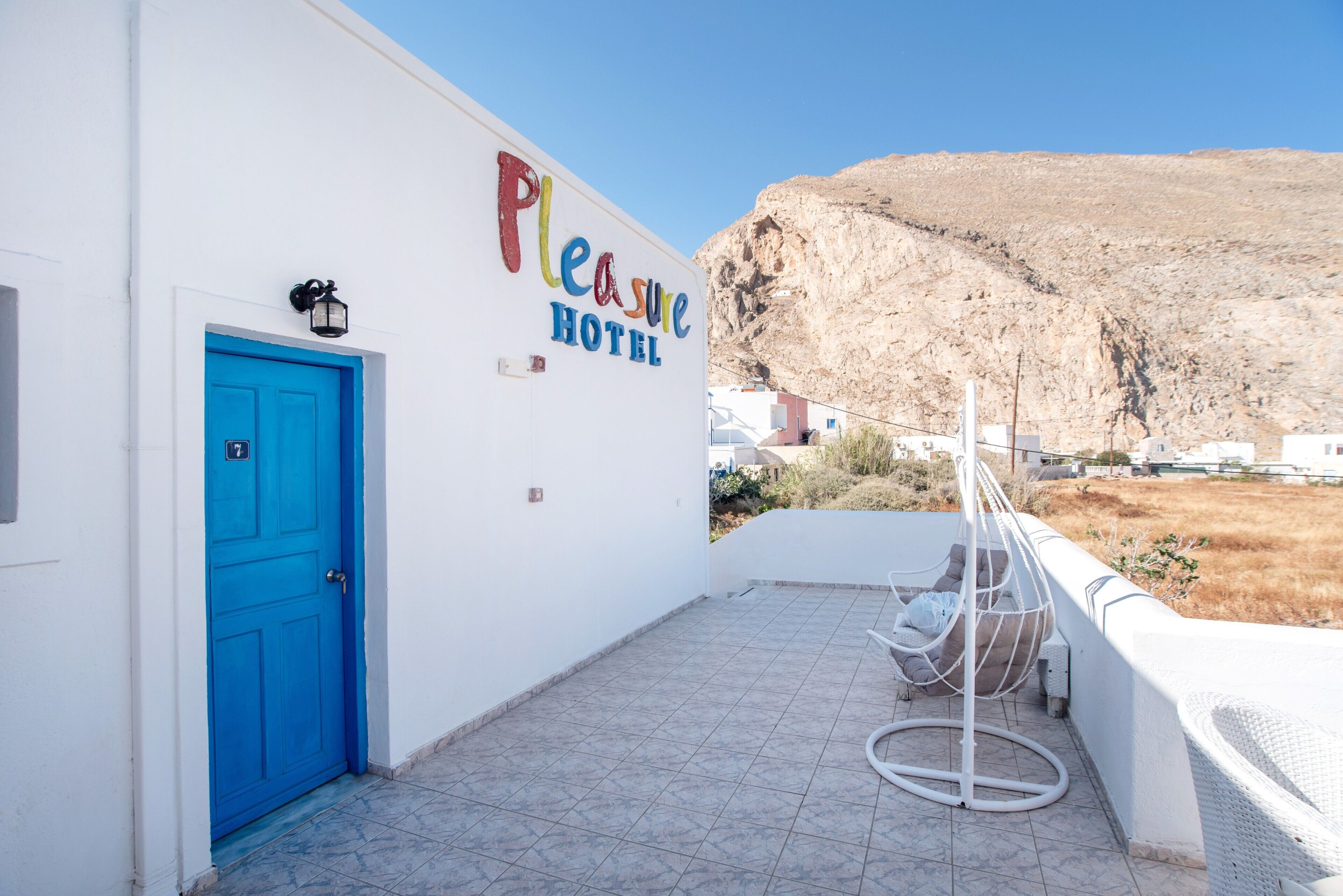 Un hotel chiamato 'Pleasure Hotel' con una porta blu, un muro esterno bianco e vista su un paesaggio montuoso.