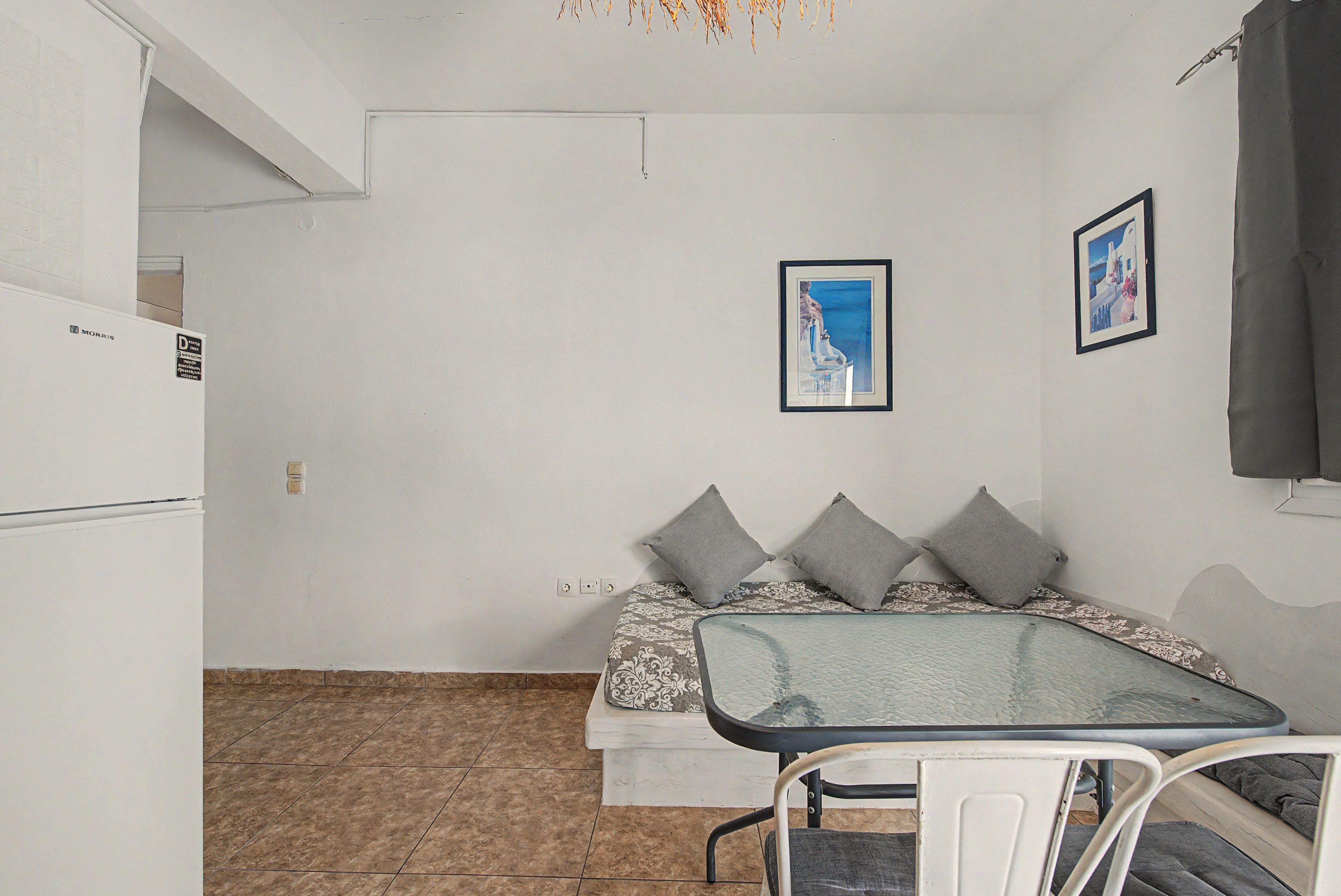Una stanza piccola e funzionale con un letto, un tavolo e un frigorifero. Due quadri appesi alle pareti.