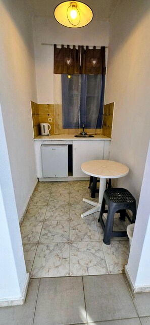A compact kitchen with a small refrigerator, a sink, a coffee maker, a table, and a stool.