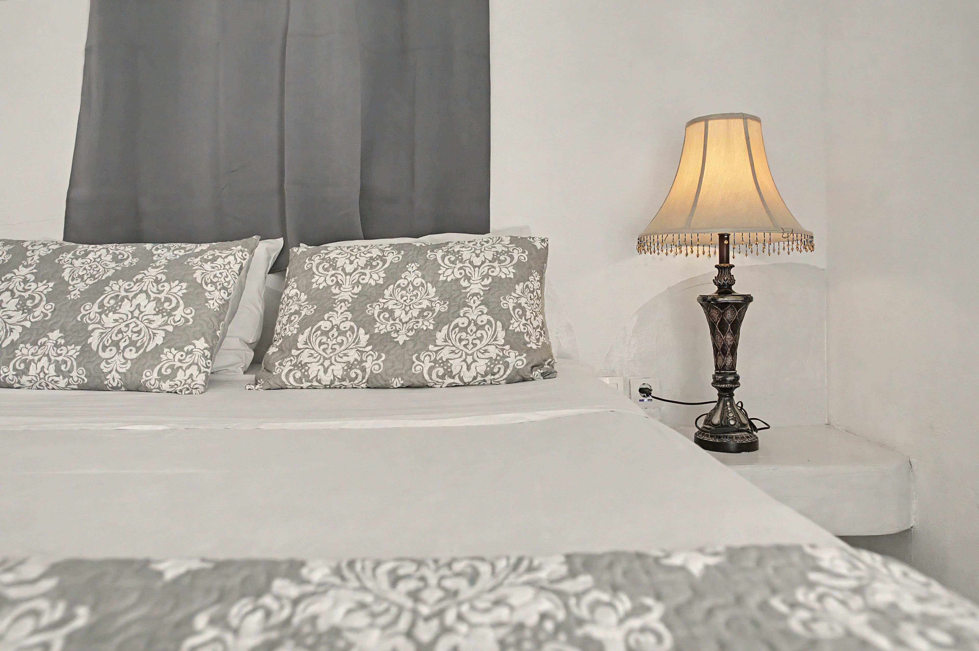 Un lit recouvert d’une couvre-lit à motifs blancs et gris, deux oreillers à motifs damassés gris et blancs, et une lampe de table avec un abat-jour beige et un pied décoratif.