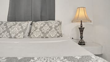 Un lit recouvert d’une couvre-lit à motifs blancs et gris, deux oreillers à motifs damassés gris et blancs, et une lampe de table avec un abat-jour beige et un pied décoratif.