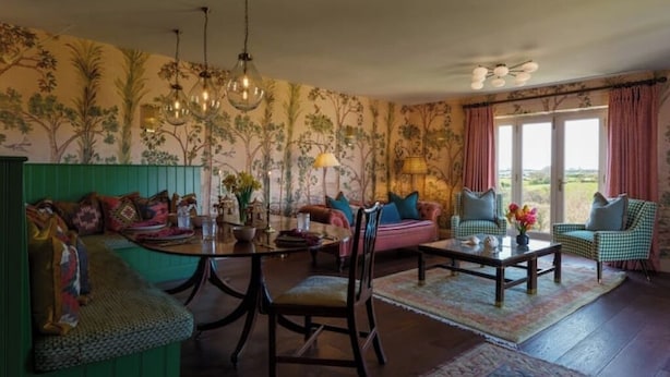 Un salon chaleureux avec un banc vert, une table ronde et un canapé. On y trouve deux fauteuils, une lampe et une vue sur l’extérieur par une fenêtre.