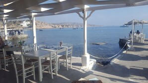 An outdoor seating area with tables and chairs overlooking a body of water, with boats visible in the distance.