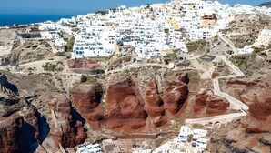 An aerial view of a coastal town with white buildings, a harbor with boats, and rocky terrain.