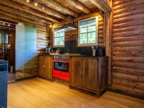 Eine Küchenzeile in einer Blockhütte mit Holzwänden, einem roten Ofen und einem Fenster mit Blick auf Grünflächen.