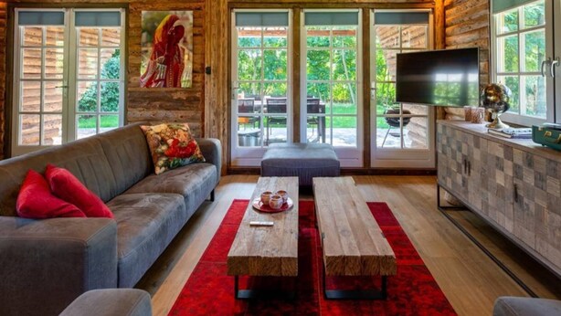 A cozy log cabin interior with a sofa, two wooden tables, a TV, and a kitchen area. - Spacious Wooden Chalet With Covered Terrace (Baarle-Nassau)