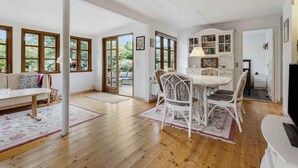 A spacious living area with a wooden floor, a dining table with chairs, and a white sofa. There are large windows with wooden frames, a bookshelf, and a door leading to an outdoor area. - 5 Star Holiday Home in Nykobing Sj (Sjællands Odde)