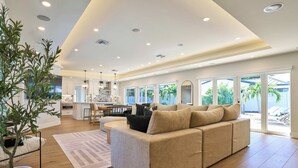 A spacious living room with a beige sectional sofa, a large potted plant, and a view of the kitchen and outdoor patio through large sliding glass doors.