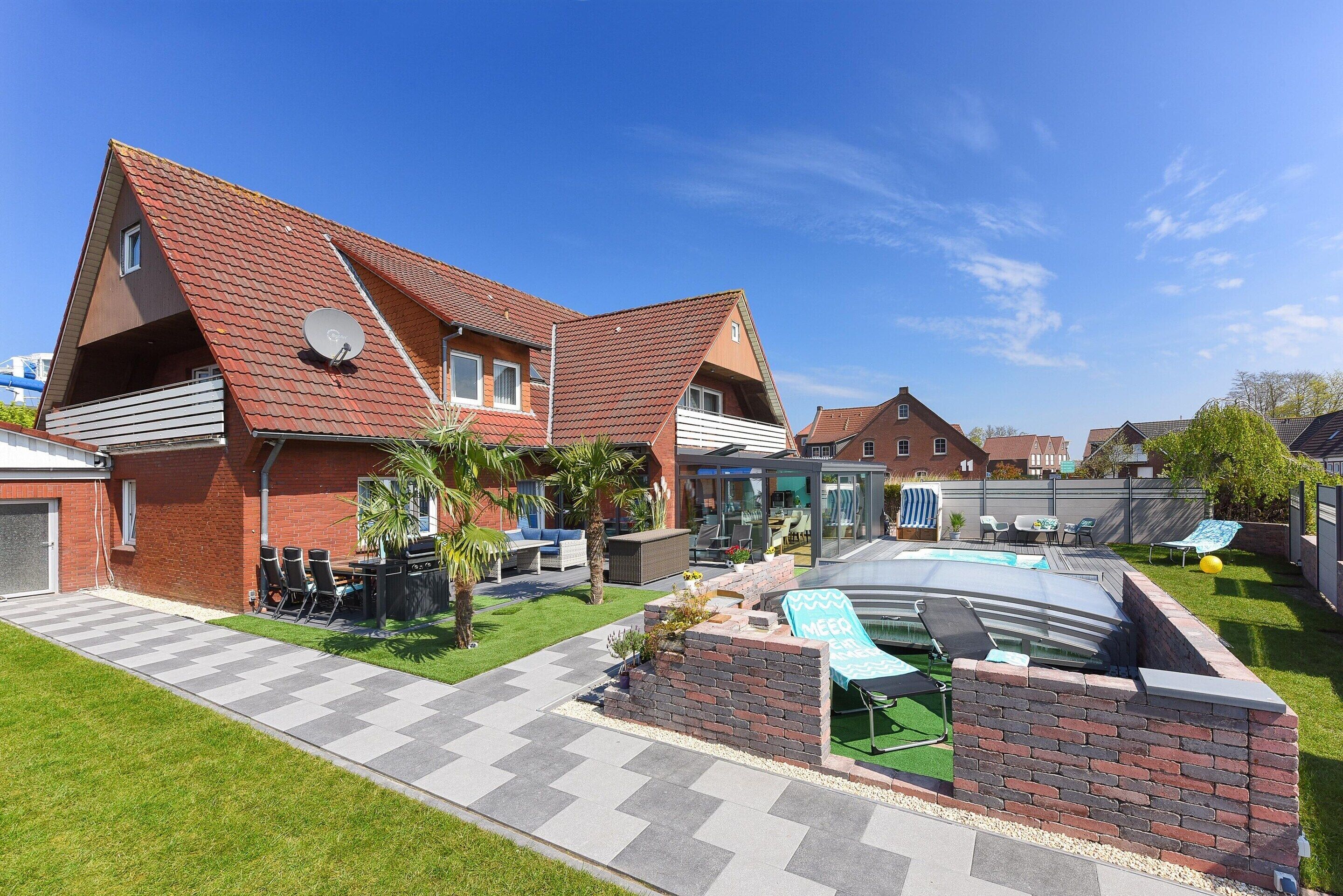 A two-story house with a swimming pool, a patio with chairs, and a brick wall.