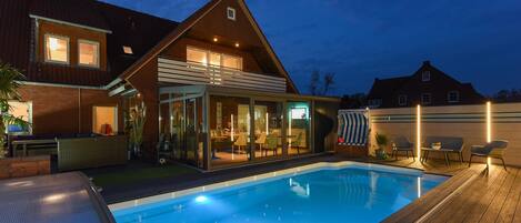 A modern house with a swimming pool, a covered patio, and a well-lit interior.