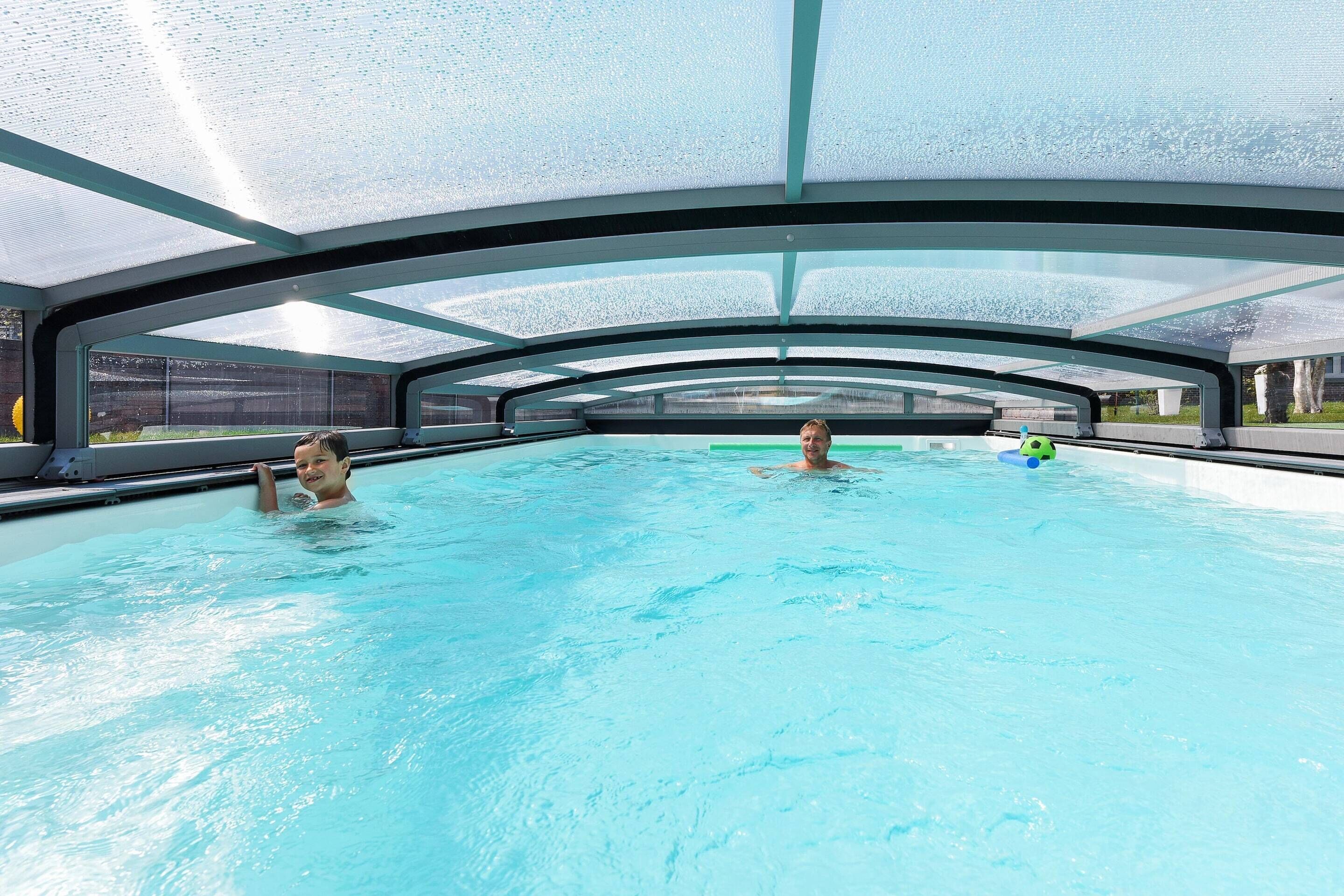 An indoor swimming pool with a glass ceiling, clear water, and a view of the outside.