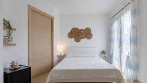 Une chambre à coucher comprenant un lit, une table de chevet, une œuvre d’art accrochée au mur, une porte et une fenêtre avec des rideaux.