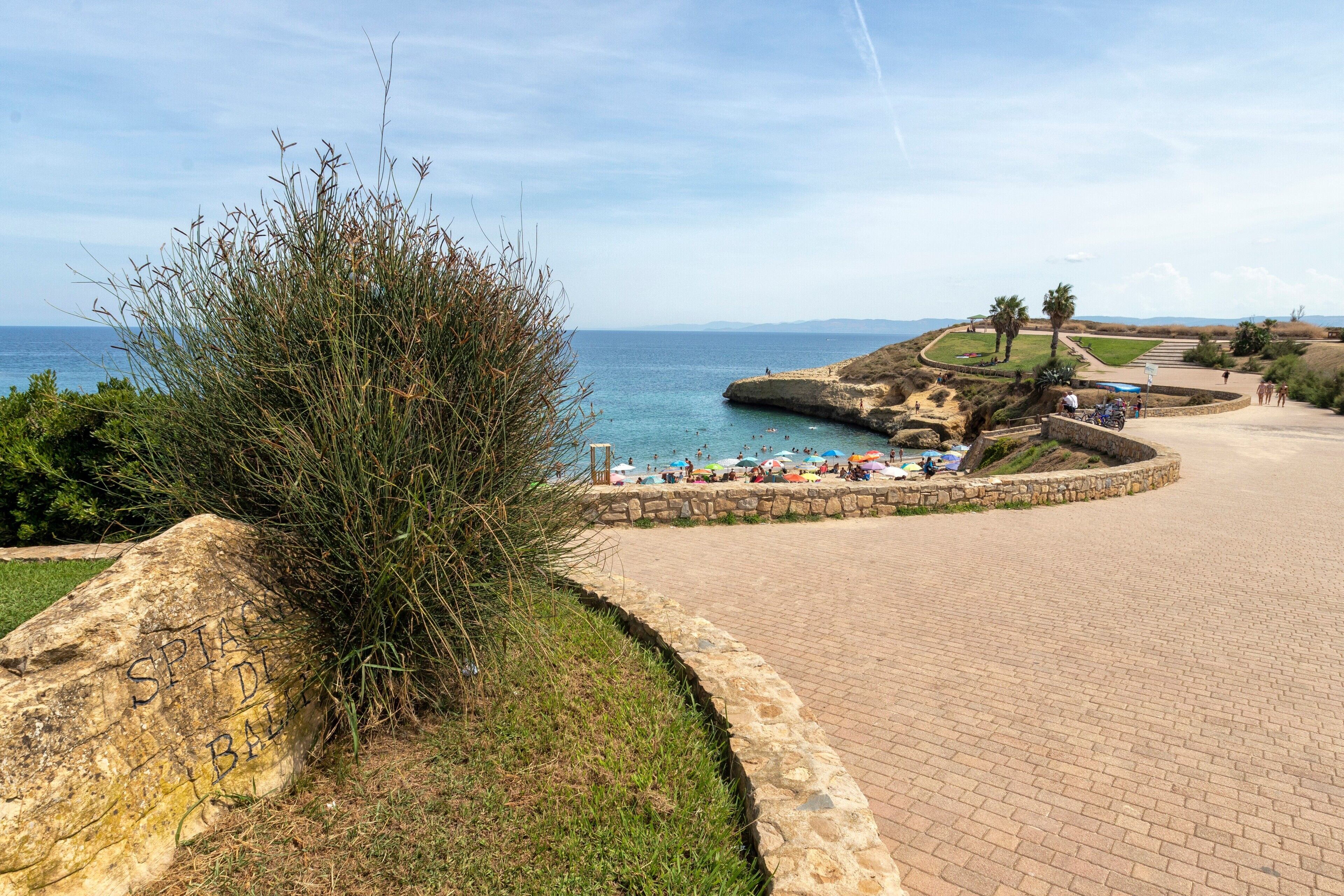 Una zona costera con un camino pavimentado, una gran roca con inscripciones y una playa con gente y sombrillas.