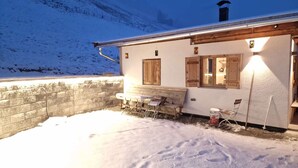 A snow-covered outdoor area with a wooden bench and a table, a stone wall, and a building with wooden shutters and a chimney. - Cabin With Sauna Near Spieljoch Ski (Fuegenberg)