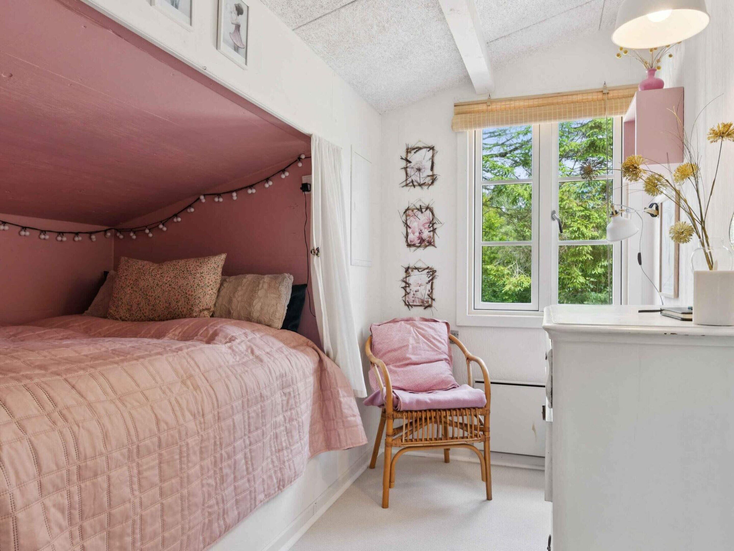 Una camera da letto con un letto rosa, una poltrona in vimini e una finestra vista alberi.