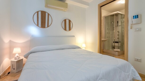 A bedroom with a bed, bedside lamps, a wall-mounted air conditioner, and a glass-enclosed shower. - Studio Apartment 'L'Angolo Monolocale- Porto Torres' with Shared Terrace, Wi-Fi and Air Conditioning (Porto Torres)