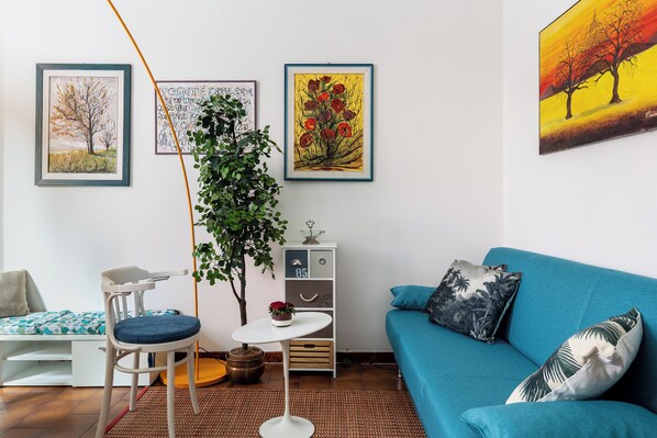 Een woonkamer met een blauwe bank, een witte bijzettafel, een stoel en een kleine plant. Er hangen drie kunstwerken aan de muur.