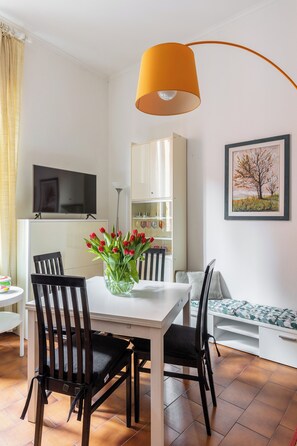 Een eethoek met een witte tafel, zwarte stoelen, een vaas rode tulpen, een gele lamp, een televisie aan de muur en een boekenkast.