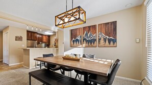 Un coin repas avec une table en bois, des chaises noires et un banc. Trois tableaux représentant des montagnes sont accrochés au mur.