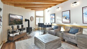 Un salon chaleureux avec un plafond en bois, une cheminée, un téléviseur et un coin repas comprenant des chaises et une table.